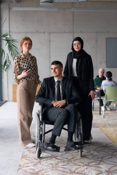 A diverse business team poses confidently, including a smiling director seated in a wheelchair, flanked by two professional female colleagues, one wearing a hijab and the other holding a tablet, in a modern office environment.