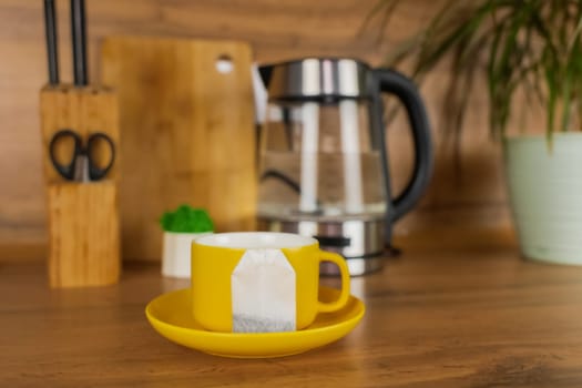 A yellow cup of tea stands on a yellow saucer and on the saucer lies a tea bag on a wooden table against the background of a teapot, a plant and kitchen utensils. The concept of tea drinking, break. High quality photo