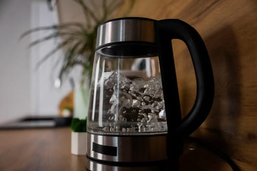 Close-up of an electric kettle with a glass base with boiling water bubbling bubbles stands on the wooden countertop of a modern kitchen, behind the kettle you can see kitchen utensils, a plant and a black sink. Boiling water, electrical appliances. High quality photo
