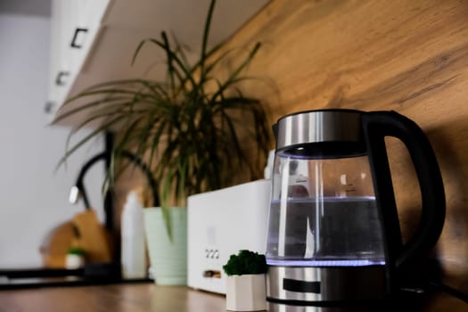 Close-up of kitchen wooden countertop with electric kettle with lighting, plants, toaster, kitchen sink, focus on kettle. Modern Kitchen, Functionality, Kitchen Appliances Concept. High quality photo