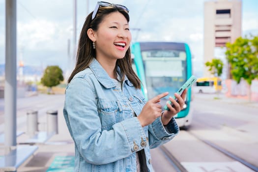 Cheerful young woman wearing a denim jacket using her smartphone while waiting at a tram stop. A modern tram is visible in the background. Bright and lively urban environment.
