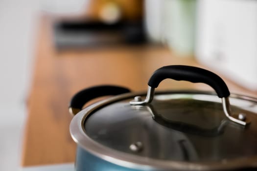 Close-up in focus: a blue saucepan with a transparent lid on which rubber pads on the handles against the background of a wooden kitchen are out of focus. High quality photo