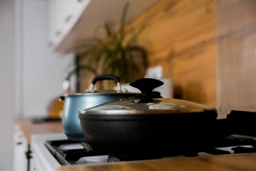 Close-up of a black frying pan with a transparent lid and a blue saucepan with a transparent lid and silicone pads on the handles against the background of a modern wooden-white kitchen out of focus. The concept of cooking, cooking, boiling and frying. High quality photo