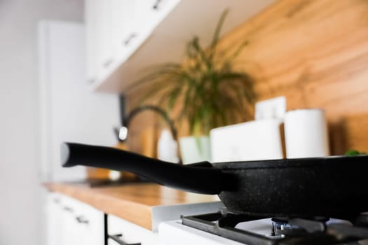 A black frying pan with a black handle stands on a white gas stove on which a fire is burning against the background of a modern wooden white kitchen in out-of-focus. The concept of frying, cooking, kitchen island. High quality photo