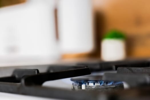 Close-up of a white gas stove with a black cast iron grate and a black burner on which a blue fire burns against the background of a wooden kitchen in out-of-focus with kitchen utensils and a plant. Gas stove, cooking concept. High quality photo