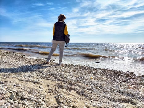 A woman stands on a rocky beach, gazing out at the shimmering water under a clear sky, embodying a sense of freedom and contemplation.