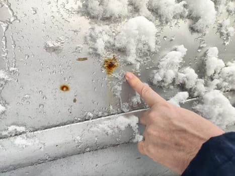 A person points at visible rust spots on a car's exterior covered with snow during cold weather in a neighborhood.