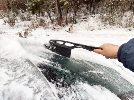 A driver clears snow off their car with a brush as cold winter weather blankets the surroundings in a snowy atmosphere.
