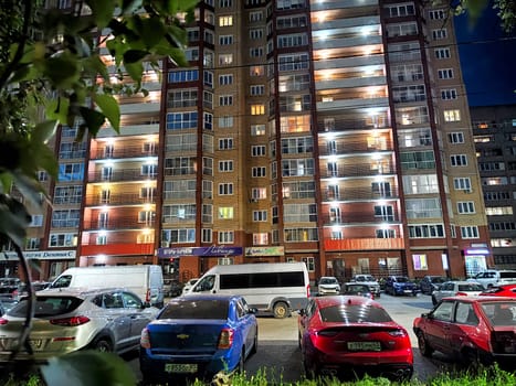 Kirov, Russia - August 11, 2023 - Apartment buildings glow with lights from balconies as parked cars fill the foreground on a bustling urban night.
