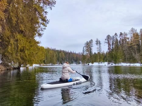 Kirov, Russia - March 14, 2024: A person enjoys paddleboarding on a calm lake, with green trees and a hint of snow in the background during early evening.