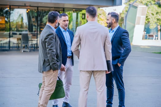 Four businessmen standing outdoors in an informal meeting, engaged in conversation. Dressed in smart-casual attire, they display collaboration and professionalism in a modern urban space.