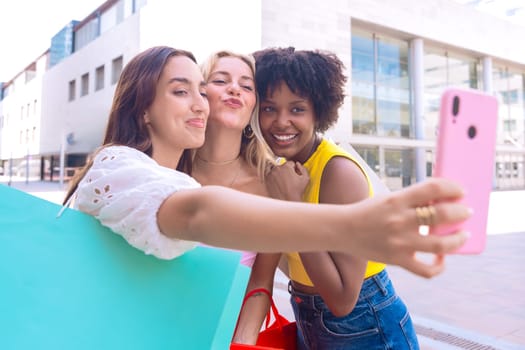 A group of three young women with diverse features smile while taking a selfie in an urban setting. They are dressed in casual summer outfits, enjoying a fun moment outdoors in the city.