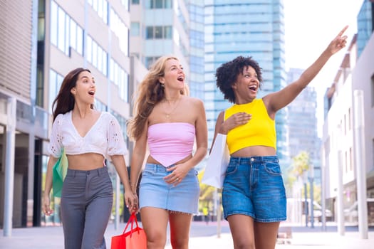 A group of three young women walk through a modern city street carrying shopping bags. One of them points excitedly at something while they all smile, enjoying a fun and lively shopping day.