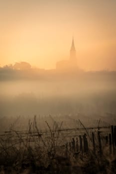 A serene vineyard landscape unfolds in Tabanac, Gironde, as the sun rises behind the hills. The winter fog envelops the vines, casting a mystical glow with warm orange tones