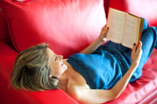 Attractive mature woman smiling in enjoyment as she reads a book while relaxing at home on a red leather couch