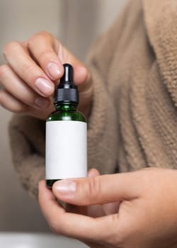 Female hands with natural manicure holding green cosmetic dropper bottle, label mockup. Close up of woman wearing light brown bathrobe using serum or essence