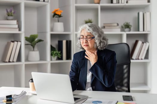An elderly businesswoman sitting in a modern home office, coughing and holding her throat, reflecting discomfort while focusing on her laptop.