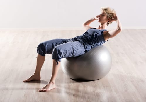 Young barefoot woman doing sit ups with hands behind head on large gray colored stability ball on hardwood floor