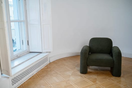 dark gray minimalist style armchair stands in the room. wooden beautiful floor. warm shade parquet lies on the floor. armchair stands against white wall. daylight from large bright window