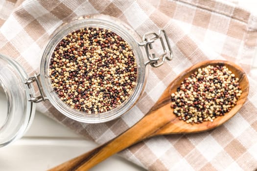 Mixed uncooked quinoa seeds in jar on a checkered napkin. Top view.