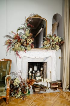 living room with fireplace. fireplace made of white stone. wooden parquet on the floor. around the fireplace a lot of decor, lots of flowers and candles on the floor, also old books with brown covers.
