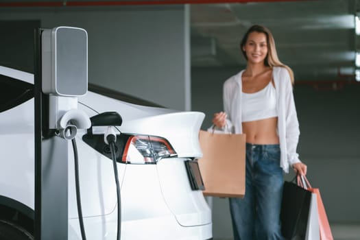 Young woman travel with EV electric car to shopping center parking lot charging in downtown city showing urban sustainability lifestyle by green clean rechargeable energy of electric vehicle innards