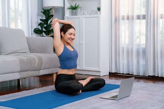 Asian woman in sportswear doing yoga exercise on fitness mat as her home workout training routine. Healthy body care lifestyle woman watching online yoga video on laptop. Vigorous