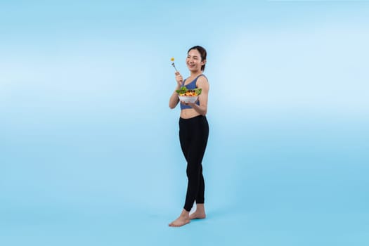 Young sporty Asian woman in sportswear holding salad bowl fill with vibrant of fruit and vegetable. Natural youthful and fit body lifestyle with balance nutrition on isolated background. Vigorous