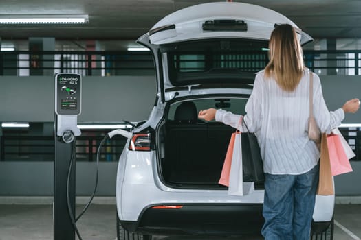 Young woman travel with EV electric car to shopping center parking lot charging in downtown city showing urban sustainability lifestyle by green clean rechargeable energy of electric vehicle innards