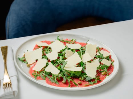 Beef Carpaccio cold appetizer with parmesan, capers and arugula on white plate in restaurant table interior