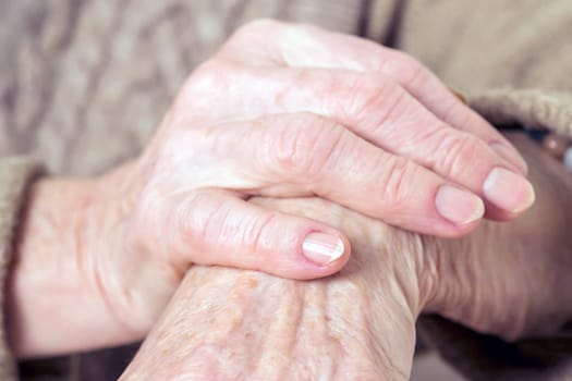 The hands of an elderly well-groomed woman in a casual sweater. The pensioner has crossed her beautiful arms with wrinkles on her skin and is sitting at the table in her cozy home.