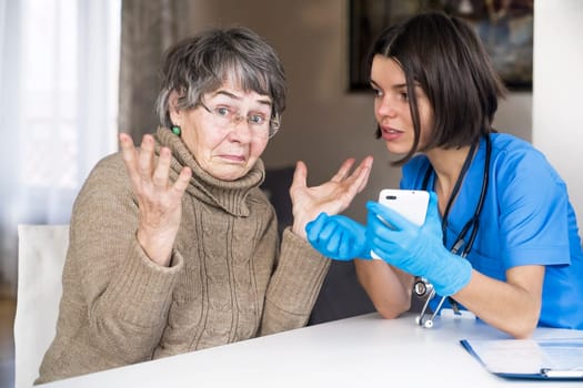 A nurse in a medical suit takes care and explains to an elderly patient how to use applications on a smartphone. Grandmother 80 years old, does not understand how to communicate with a doctor online.