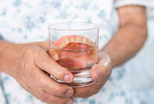 Asian senior woman patient holding and washing denture in water cleanser glass for good chewing.