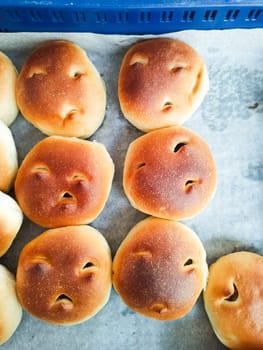 Unique buns with facial expressions are arranged in a bakery setting. The freshly baked goods draw attention with their playful designs, inviting customers to explore the treats.