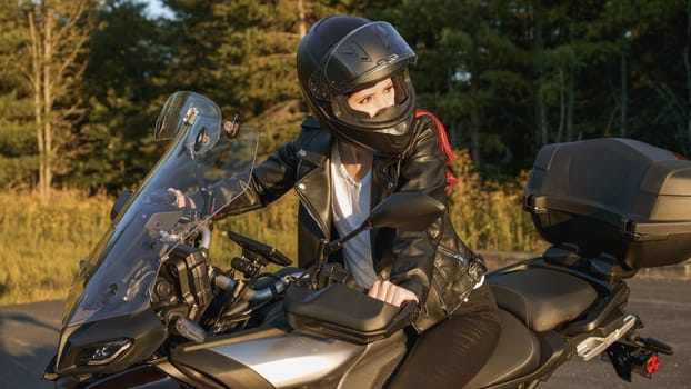 Young biker girl riding touring motorbike on forest road. Beautiful woman with red hair in leather jacket sitting on motorcycle in the park