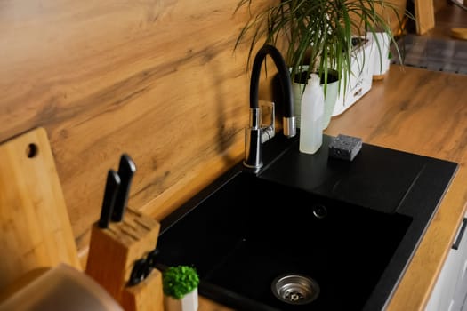 Side view of the kitchen black granite sink on with flexible black faucet on the background of wooden countertop and kitchen utensils and plants. High quality photo