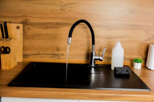Kitchen black granite sink with flexible black faucet from which water pours against the background of wooden countertop and kitchen utensils and plants. Kitchen island, interior, dishwashing concept. High quality photo