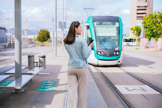 Young woman in a denim jacket standing at a tram stop, holding her smartphone while waiting for the approaching tram. Modern urban transportation and commuting concept in a city setting.