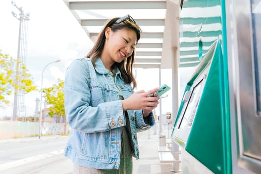 Cheerful young woman in a denim jacket using a ticket machine at a tram stop while holding her smartphone. She interacts with the touchscreen in an urban setting, preparing for her journey.