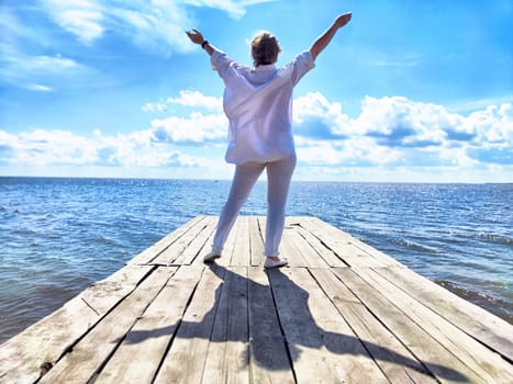 Middle-aged blonde traveler celebrates her journey on a pier, arms raised, embracing the beautiful seaside.
