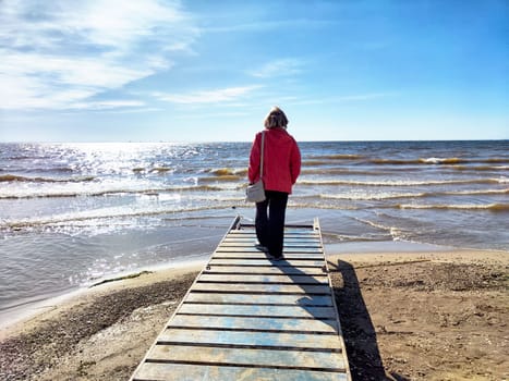 A woman strolls on a wooden boardwalk by the beach, enjoying the tranquility of the waves under a clear blue sky.