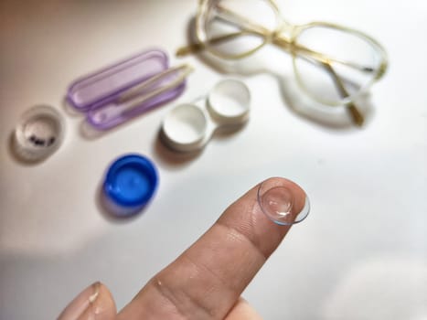 A finger holds a contact lens close to the camera while various lens care items are arranged in the background.