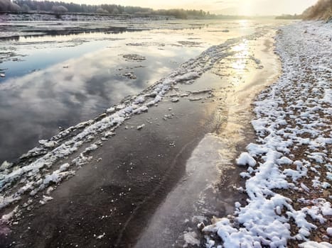 Spring thaw creates ice drift on the river as tranquil waters reflect the colorful sky and surrounding landscapes during sunset.