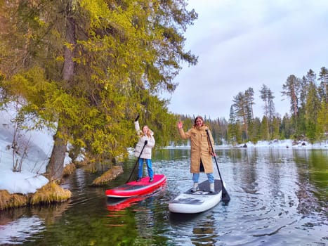 Kirov, Russia - March 14, 2024: Friends enjoy paddle boarding on a calm river, surrounded by snow-covered trees and a peaceful landscape in early spring.