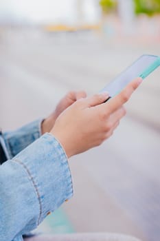 Close-up of a person wearing a denim jacket holding and using a smartphone in an urban environment. The image represents digital communication, technology, and modern connectivity.