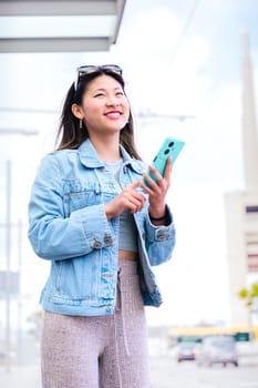 Cheerful young woman in a denim jacket holding her smartphone while looking up with a happy expression. Modern urban setting, representing digital connectivity and city lifestyle.