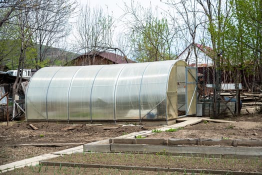 A polycarbonate greenhouse stands in a spring garden surrounded by bare trees, wooden garden beds, and freshly planted seedlings, representing early gardening efforts in March