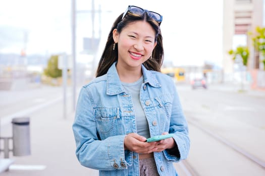 Cheerful young woman in a denim jacket holding her smartphone while looking up with a happy expression. Modern urban setting, representing digital connectivity and city lifestyle.
