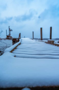 Snow-covered wooden dock extending into a calm winter lake under an overcast sky. Frosty and serene atmosphere in a natural, rustic setting. No people, peaceful scene