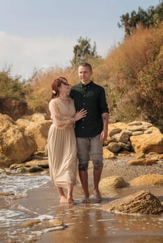 lovers and romantic newlyweds walk along the seashore. they hug each other by the waist. walk along the seashore barefoot, sea water wet their clothes. girl in beige dress looks at guy. around rocks.